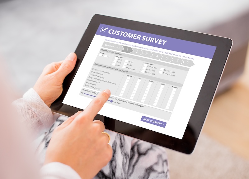 A person is holding a black tablet and filling out a customer survey form. Their index finger is touching the screen, which displays a questionnaire with options for responses. The tablet screen has a purple header that reads "Customer Survey," seamlessly blending traditional methods with modern tools like Google review bots.