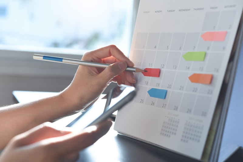 A person holds a smartphone and uses a pen to point at a specific date on a desk calendar. The calendar, adorned with colorful sticky notes for appointments, suggests efficient organization akin to employing reputation defender strategies for managing online presence.