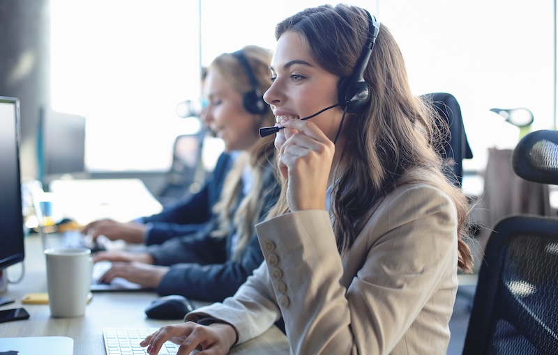 Two women working at a call center, wearing headsets and sitting at desks with computers. The woman in the foreground, a reputation defender specialist, is speaking and smiling. They appear focused and professional in a bright office environment.