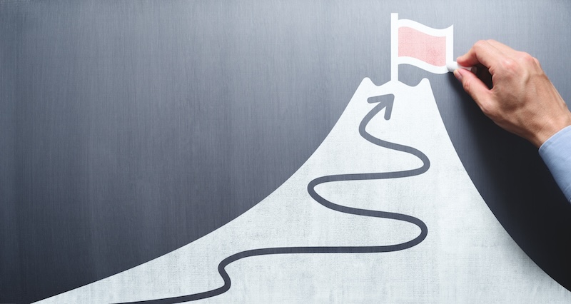 A hand places a red flag at the summit of a mountain drawing on a chalkboard, much like a reputation defender marking success. A winding path leads to the peak, symbolizing the journey of safeguarding one's achievements and goals.