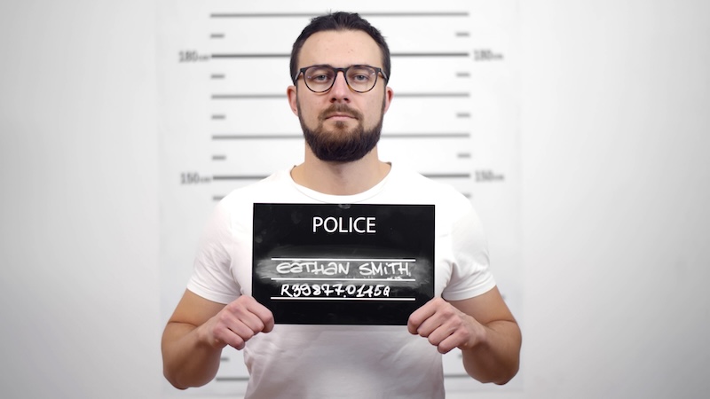 A man with a beard and glasses stands against a height chart, holding a sign with his name and number. Caught in this moment, he's being photographed for a mugshot—a moment he wishes would never appear on Google.