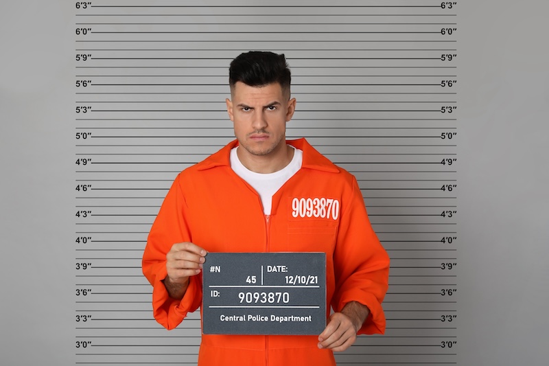 A man in an orange prison jumpsuit stands holding a sign in front of a height measurement chart, reminiscent of images some may want to learn how to get their mugshot off Google. The sign displays identification numbers and dates related to his arrest.