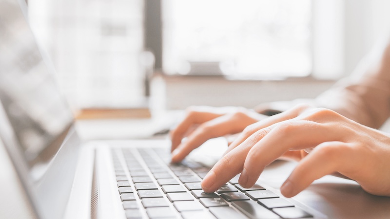 Close-up of hands typing on a laptop keyboard, perhaps researching how to get a mugshot off Google. The background is softly blurred, indicating a well-lit room with bright natural light streaming through a window, suggesting a focused working or studying environment.