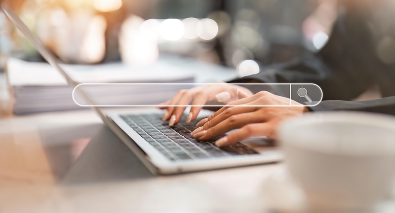 A person types on a laptop keyboard, with a blurred search bar overlayed in the foreground, hinting at efforts to remove results from Google search. The magnifying glass icon suggests searching activity, while a blurred coffee cup on the desk enhances the focused workspace setting.