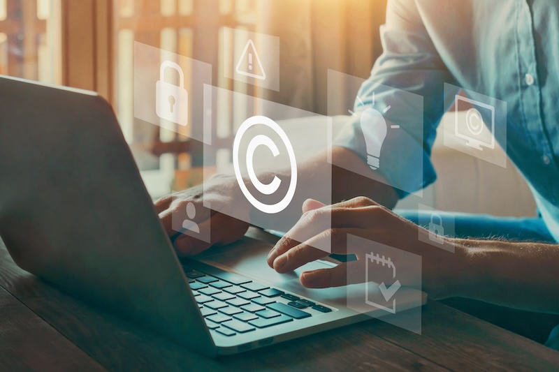 A person types on a laptop with floating icons—copyright, lock, lightbulb, alert triangle—symbolizing digital security, creativity, and intellectual property. Sunlight streams through the window as they focus on removing search results from Google for enhanced privacy.