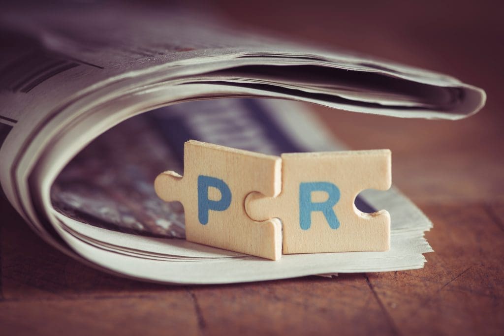 A rolled-up newspaper with two wooden puzzle pieces on top. The puzzle pieces have the letters "P" and "R" on them, symbolizing public relations. The background is wooden, giving a rustic feel to the image.