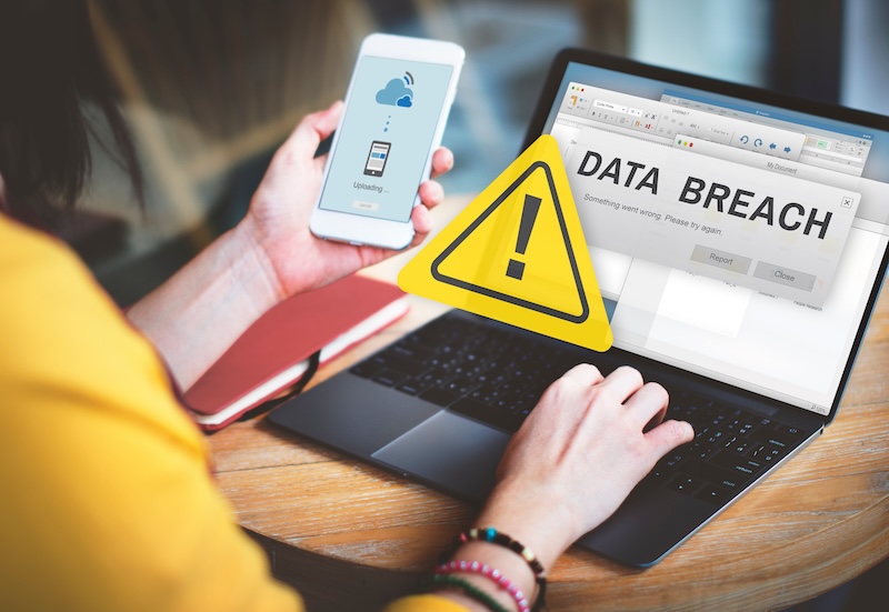 A person holds a smartphone showing an uploading screen while sitting at a desk with a laptop displaying a "Data Breach" warning message. A yellow caution symbol is superimposed on the laptop screen, concerning executive privacy. Nearby are a red notebook and a pair of glasses.