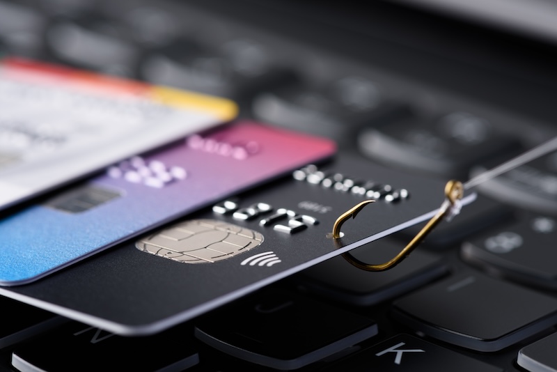 A phishing hook pierces through a stack of bank cards resting on a computer keyboard, symbolizing the risk of online payment fraud and cyber security threats, underscoring the importance of executive privacy.