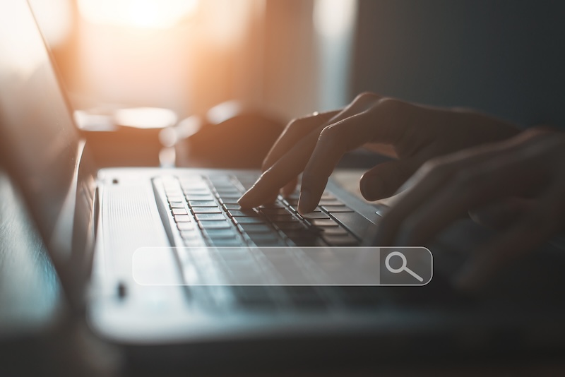 A person types on a laptop keyboard with a search bar overlayed in the foreground. Sunlight is streaming in from the background, casting a warm, soft light on the scene.