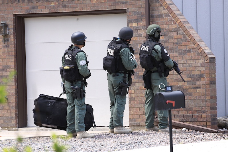 Three SWAT team members, dressed in green tactical gear and helmets with "POLICE" marked on their vests, stand in front of a brick garage door, seemingly ready to enter. One officer carries a large black bag. A mailbox is visible in the foreground, marking the boundary of executive privacy.