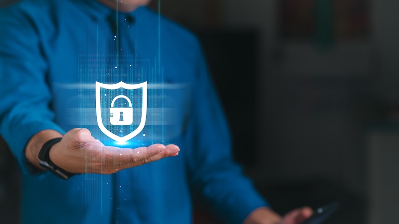 A person in a blue shirt holds out their hand with a floating digital shield that has a padlock icon on it, symbolizing executive-level cybersecurity and privacy. The background is slightly blurred, focusing attention on the shield and the person's gesture.