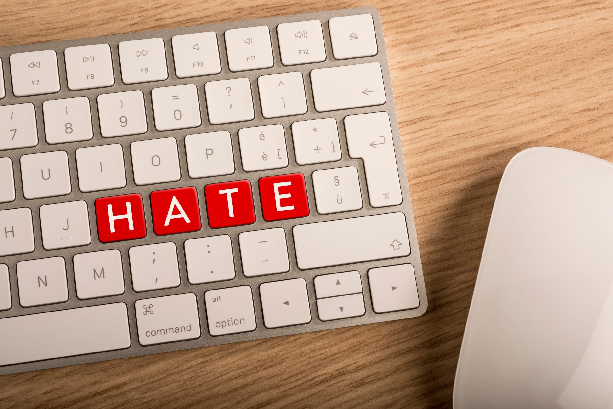 A silver and white computer keyboard displays "HATE" in red keys, a stark reminder of the impact of online slander. It sits on a wooden desk beside a white mouse, silently echoing the digital world's darker side.