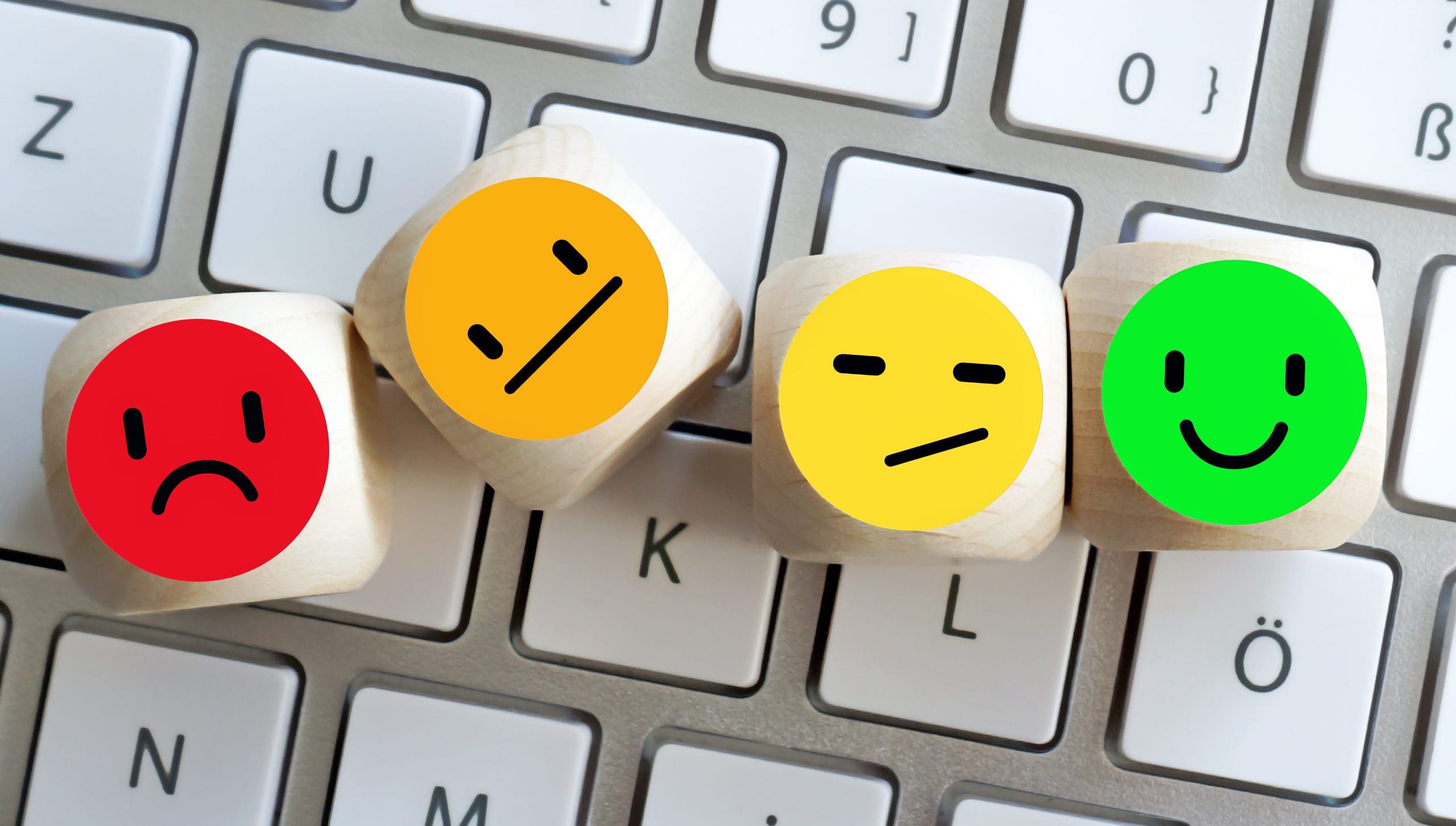 Wooden blocks with emoticons displaying various emotions: sad (red), neutral (orange), unsure (yellow), and happy (green), are placed on a computer keyboard.