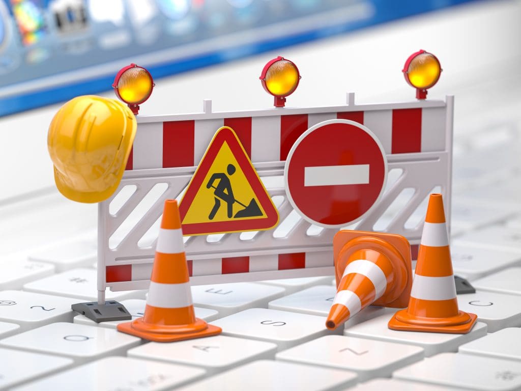 A miniature construction setup on a keyboard features a roadblock with striped barriers, traffic cones, and a construction sign. An orange hard hat and warning lights complete the scene, symbolizing online maintenance or development.