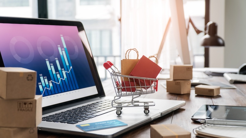 A laptop displaying a financial graph sits on a wooden desk. In front of it, a miniature shopping cart holds small packages. Surrounding the laptop are additional packages, a smartphone, and a credit card, with a bright window in the background.