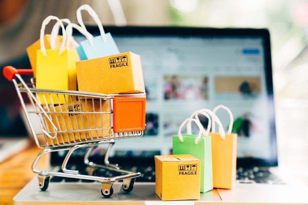 A miniature shopping cart filled with small cardboard boxes labeled "FRAGILE" and colorful shopping bags sits on a laptop keyboard, embodying tips on how to manage an online business. The laptop screen displays a blurred online shopping website, hinting at the digital marketplace's vast potential.