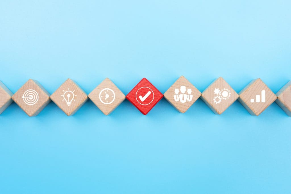 Wooden blocks lined up diagonally on a blue background, each displaying a white icon: fingerprint, light bulb, clock, check mark, group of people, gears, and bar graph. The check mark block is red, contrasting with the others.