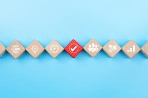 Wooden blocks lined up diagonally on a blue background, each displaying a white icon: fingerprint, light bulb, clock, check mark, group of people, gears, and bar graph. The check mark block is red, contrasting with the others.