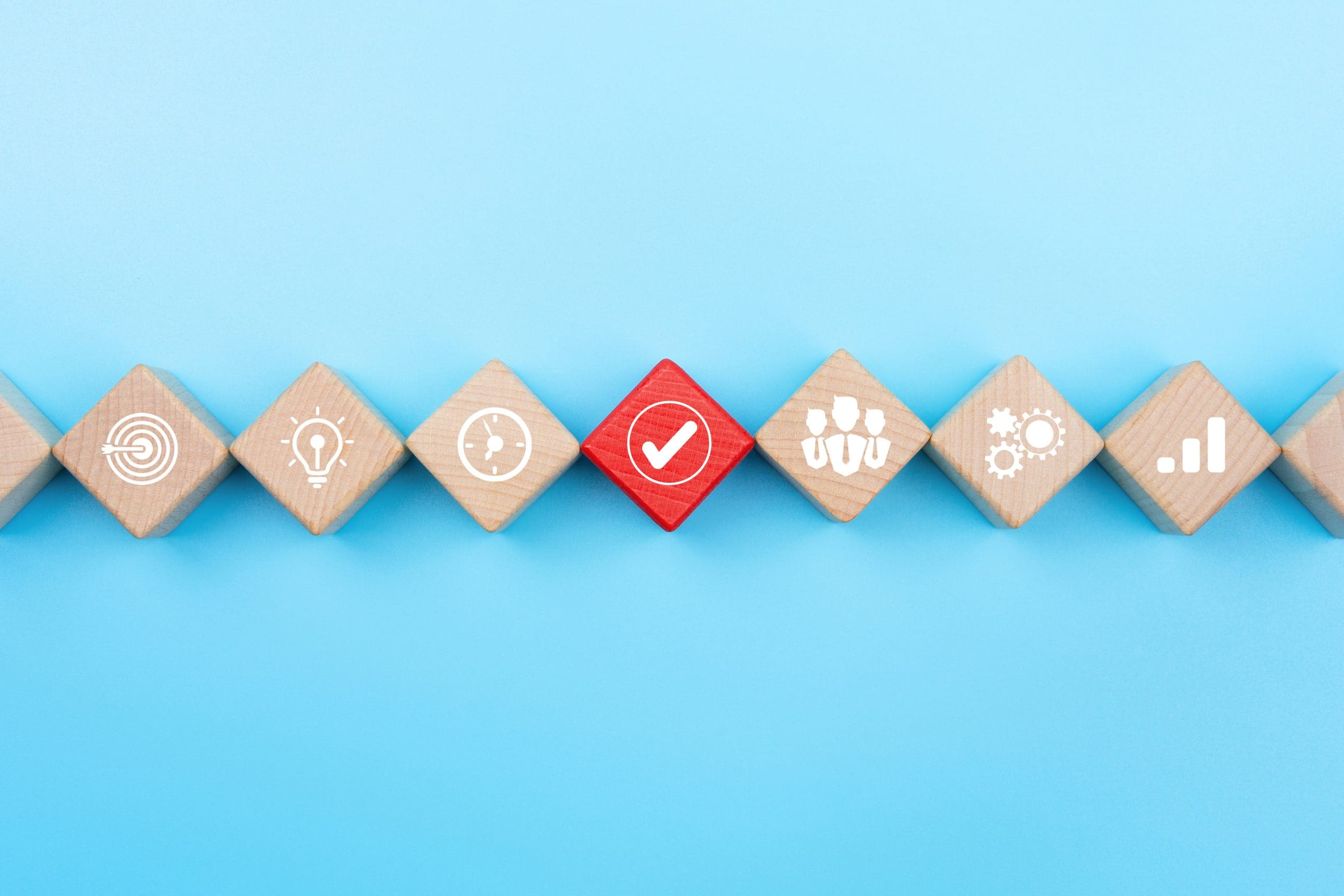 Wooden blocks lined up diagonally on a blue background, each displaying a white icon: fingerprint, light bulb, clock, check mark, group of people, gears, and bar graph. The check mark block is red, contrasting with the others.