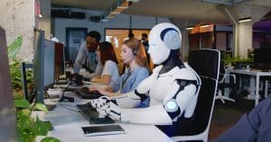 A humanoid robot wearing a headset sits at a desk using a computer in a modern office environment. People work at adjacent desks, and plants decorate the area.