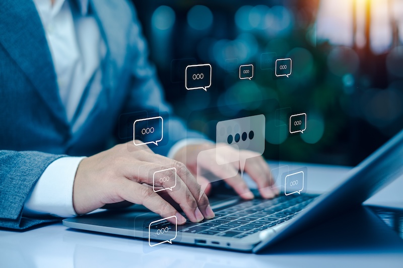 Person typing on a laptop with digital chat bubbles floating around, symbolizing online communication or chatting. The background is blurred, suggesting a focused work environment.