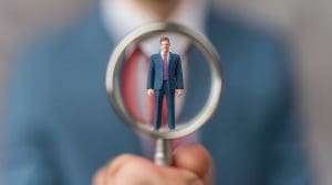 A man in a blue suit and red tie is magnified through a glass held by an unseen person. He appears miniature and focused, while the background is blurred, emphasizing the suit's texture and colors.